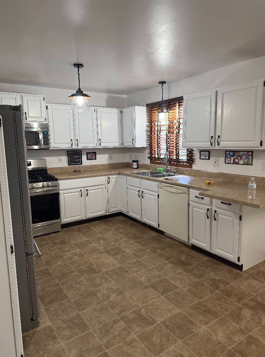 Bright kitchen with white cabinets, tan countertops, brown tile floor, and window curtains.