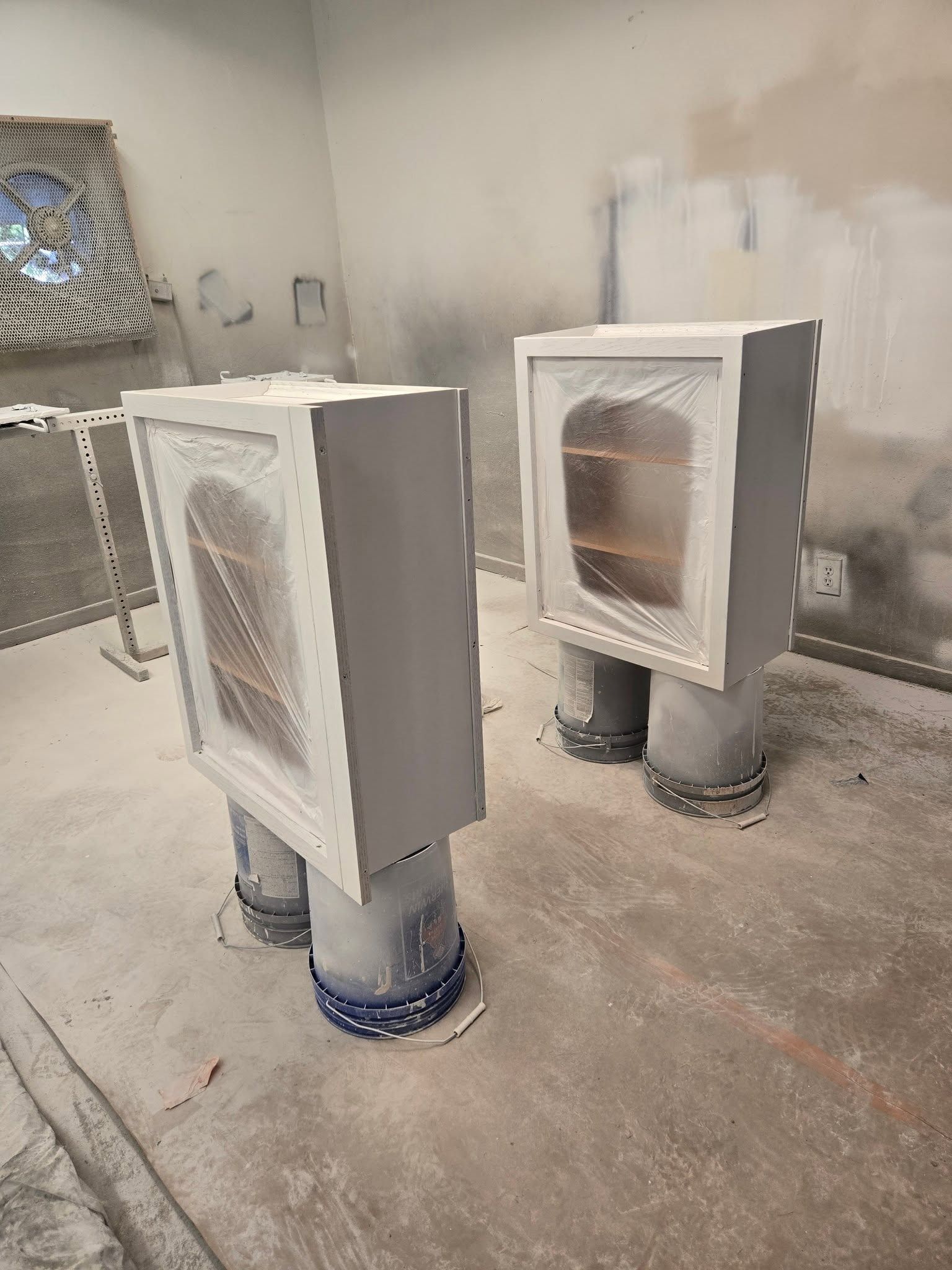 Two white vented metal cabinets on pipes in a concrete industrial room, with a metal mesh barrier behind them.