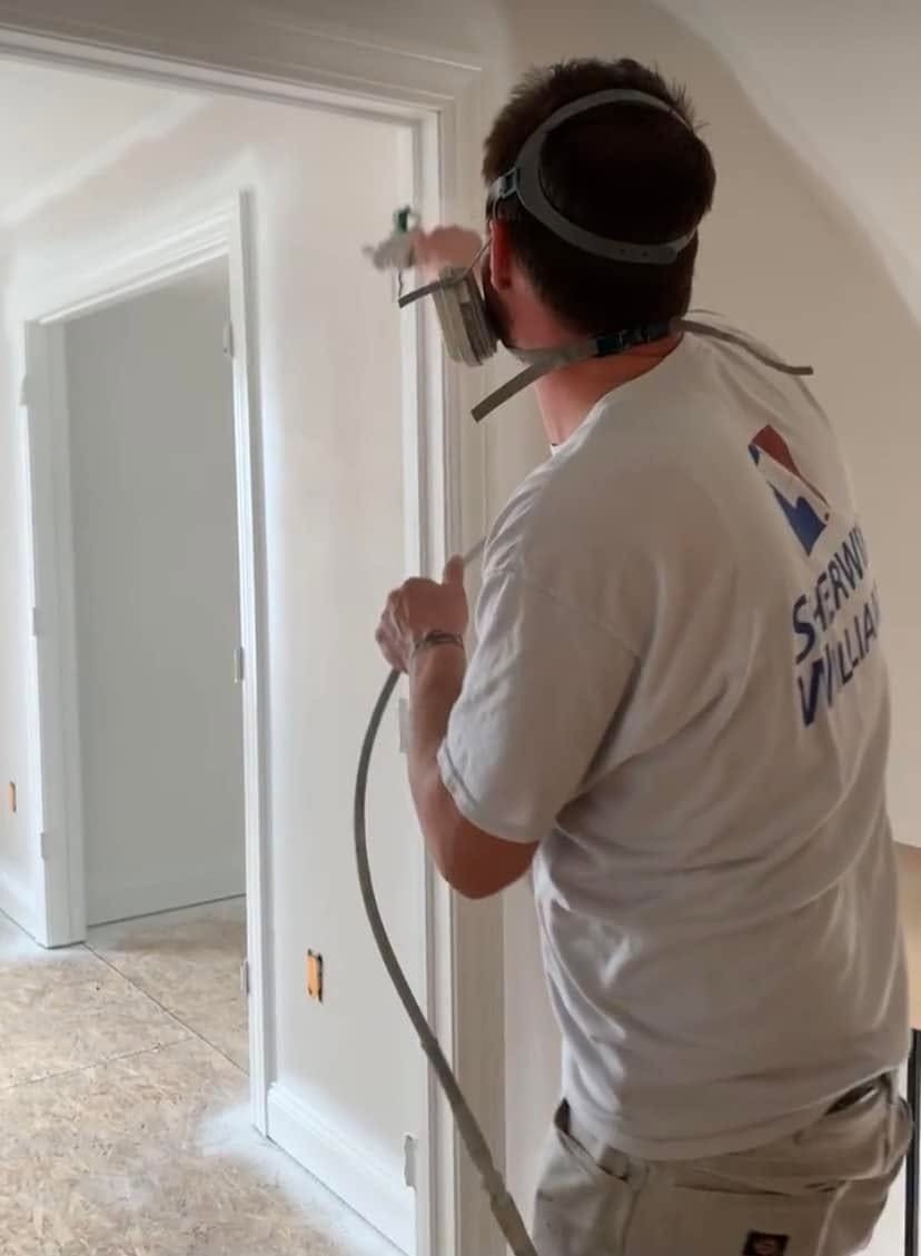 Worker spraying white paint on a doorway inside a room under renovation