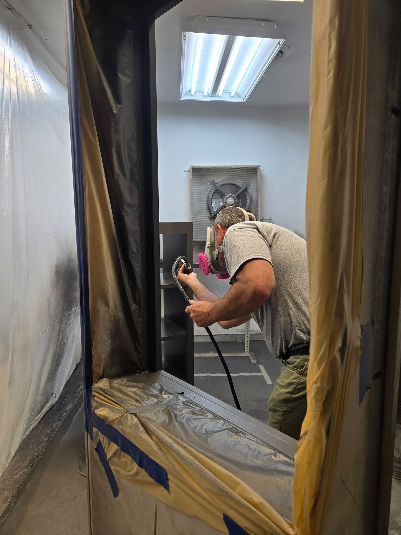 Person painting a room interior with plastic-covered walls and taped trim under a fluorescent ceiling light