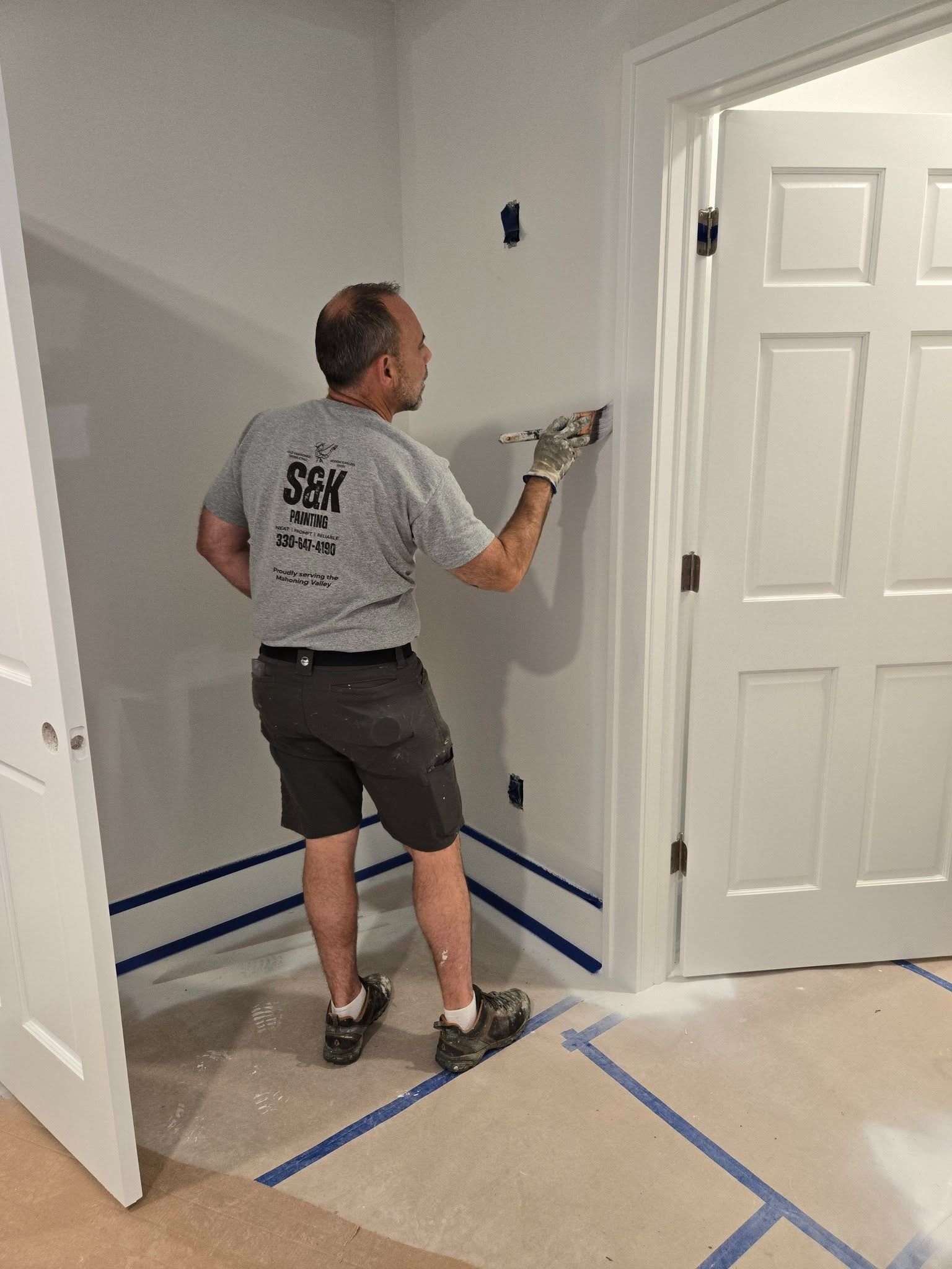 Person painting a gray wall in a room under renovation, with blue tape marking the floor.