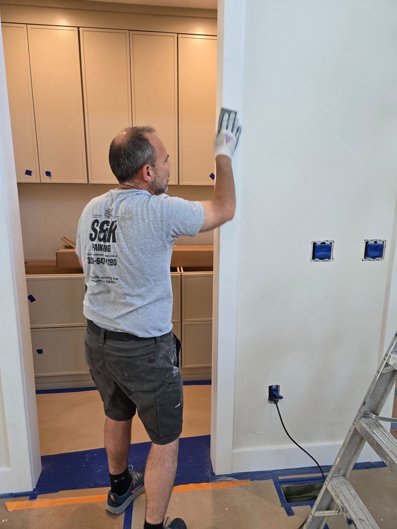Worker painting a wall in a kitchen doorway, with cabinets and a ladder nearby.