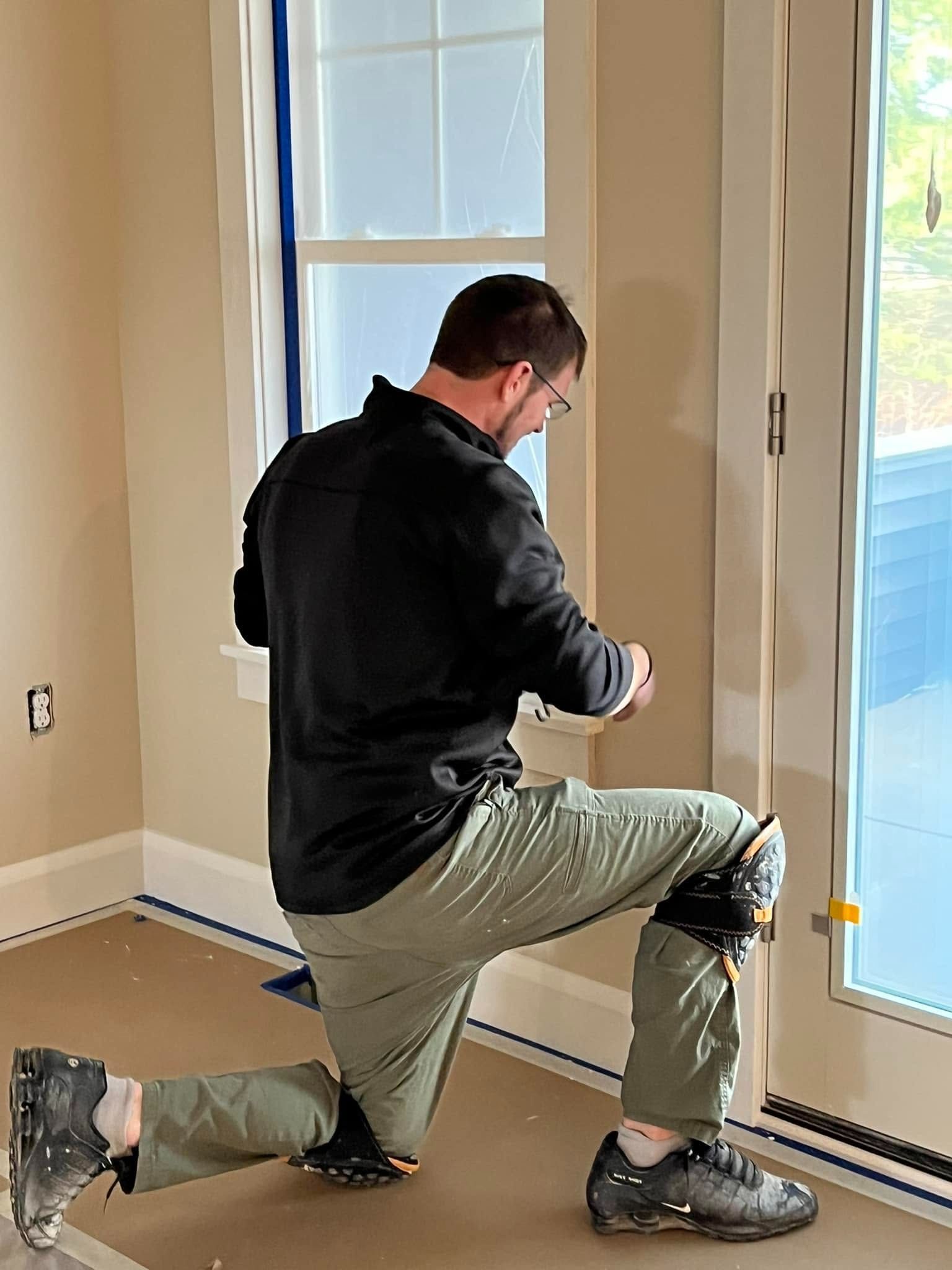 Man kneeling by a glass door, measuring or adjusting a door threshold in a bright room.