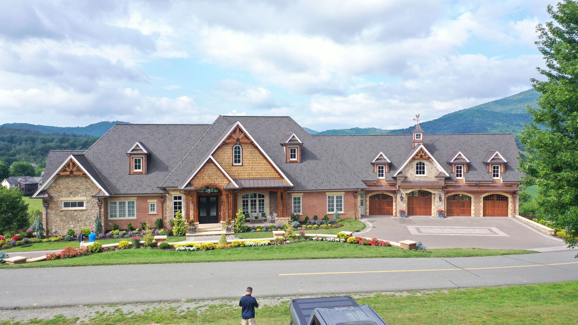 Large stone and wood home with a three-car garage, set against a backdrop of green hills and a cloudy sky.