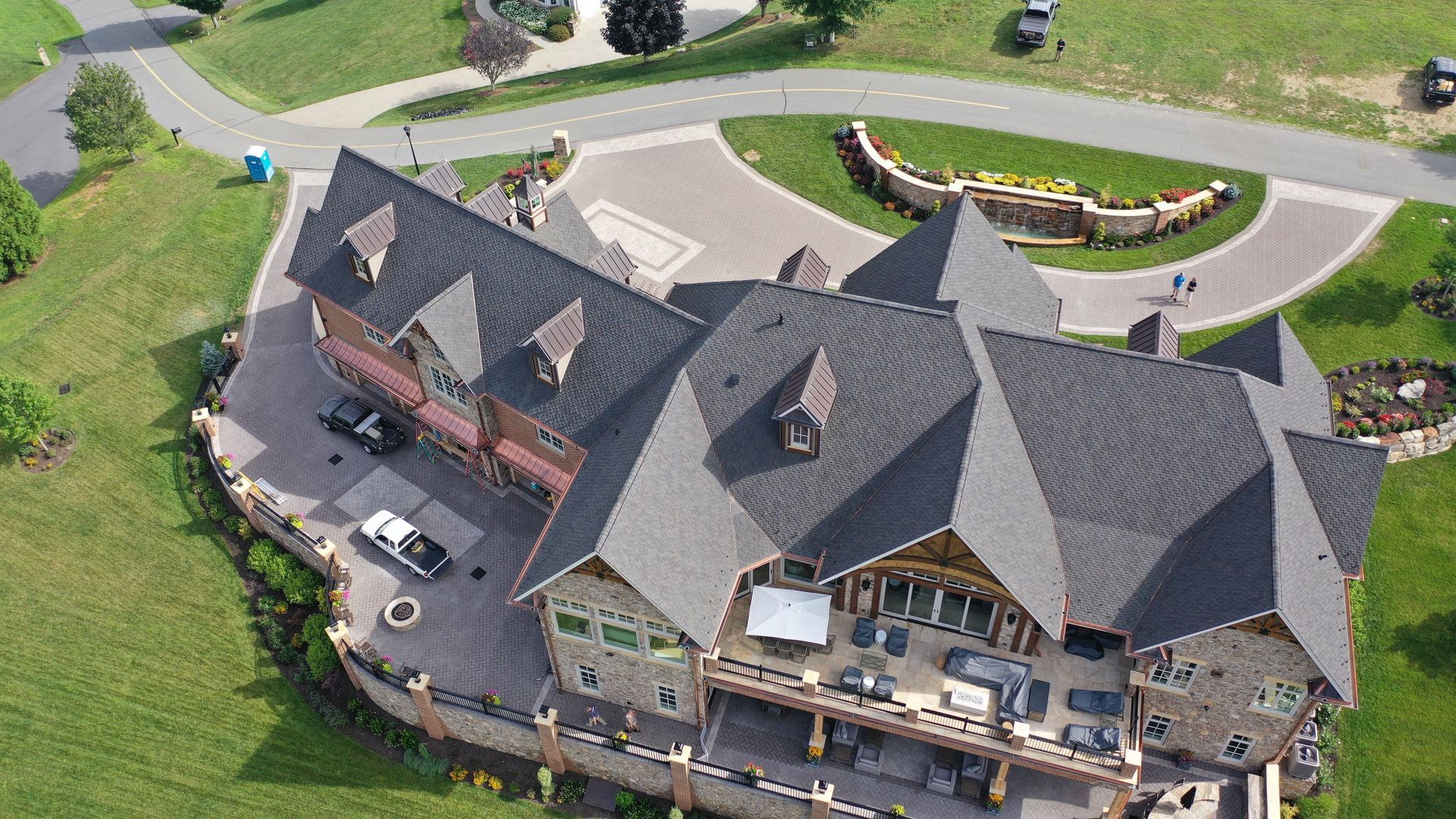 Large stone and wood mansion with gray roof, circular drive, surrounded by green grass and a winding road.