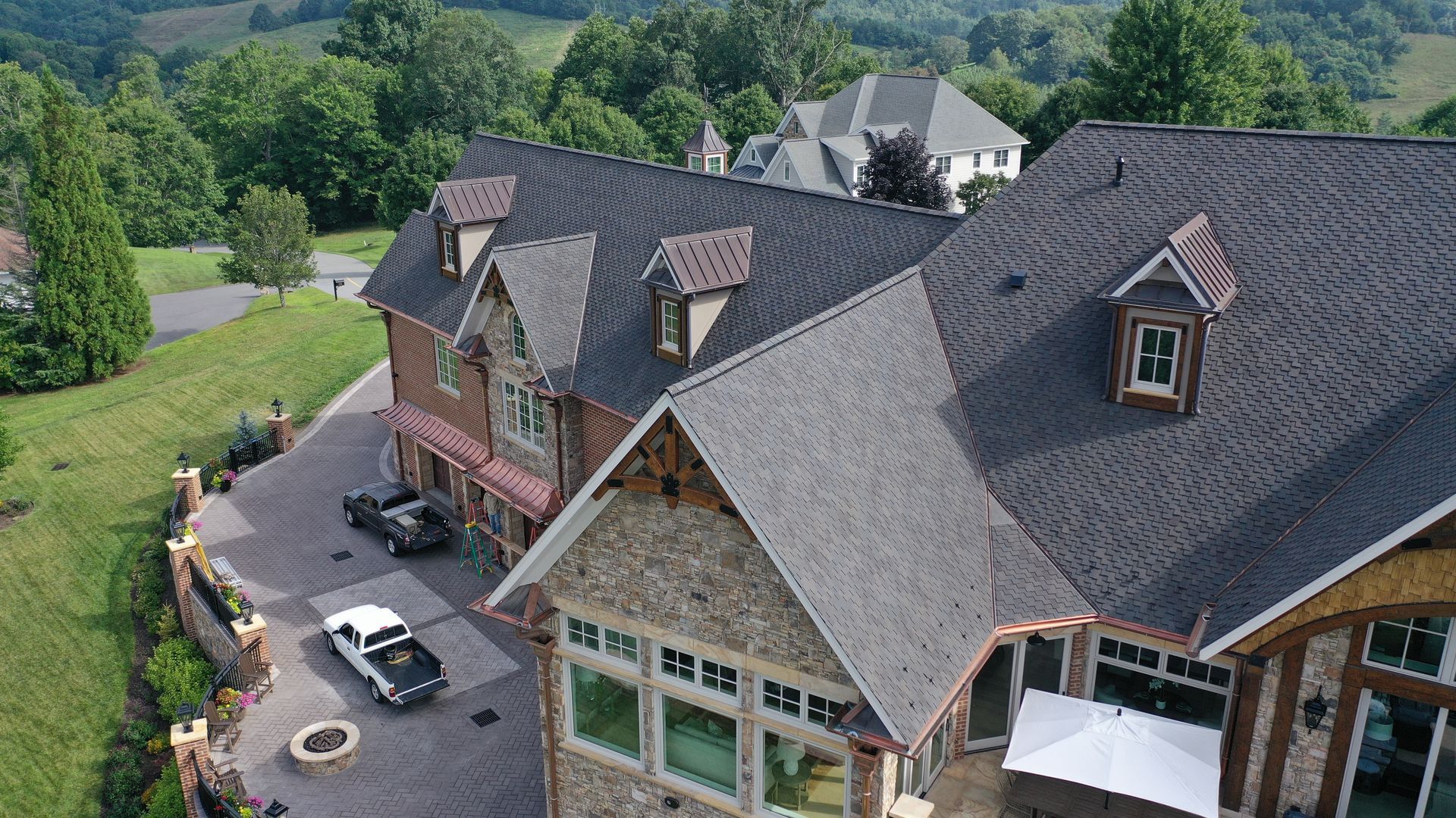 Large multi-story house with a stone and wood facade, surrounded by trees and hills. Cars parked in the driveway.