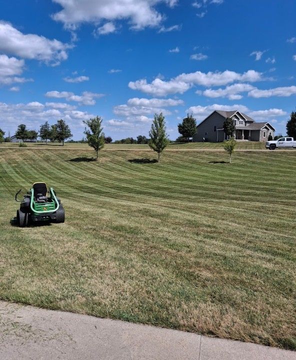 Lawn mower on a striped grassy hillside near a house under a blue sky with clouds