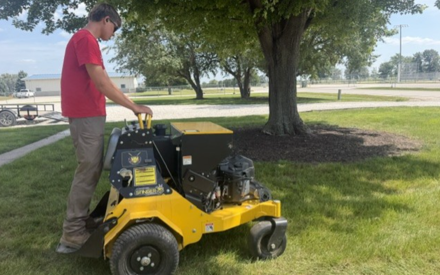 Man standing on a yellow lawn mower in a grassy park near a large tree, with a fountain in the background