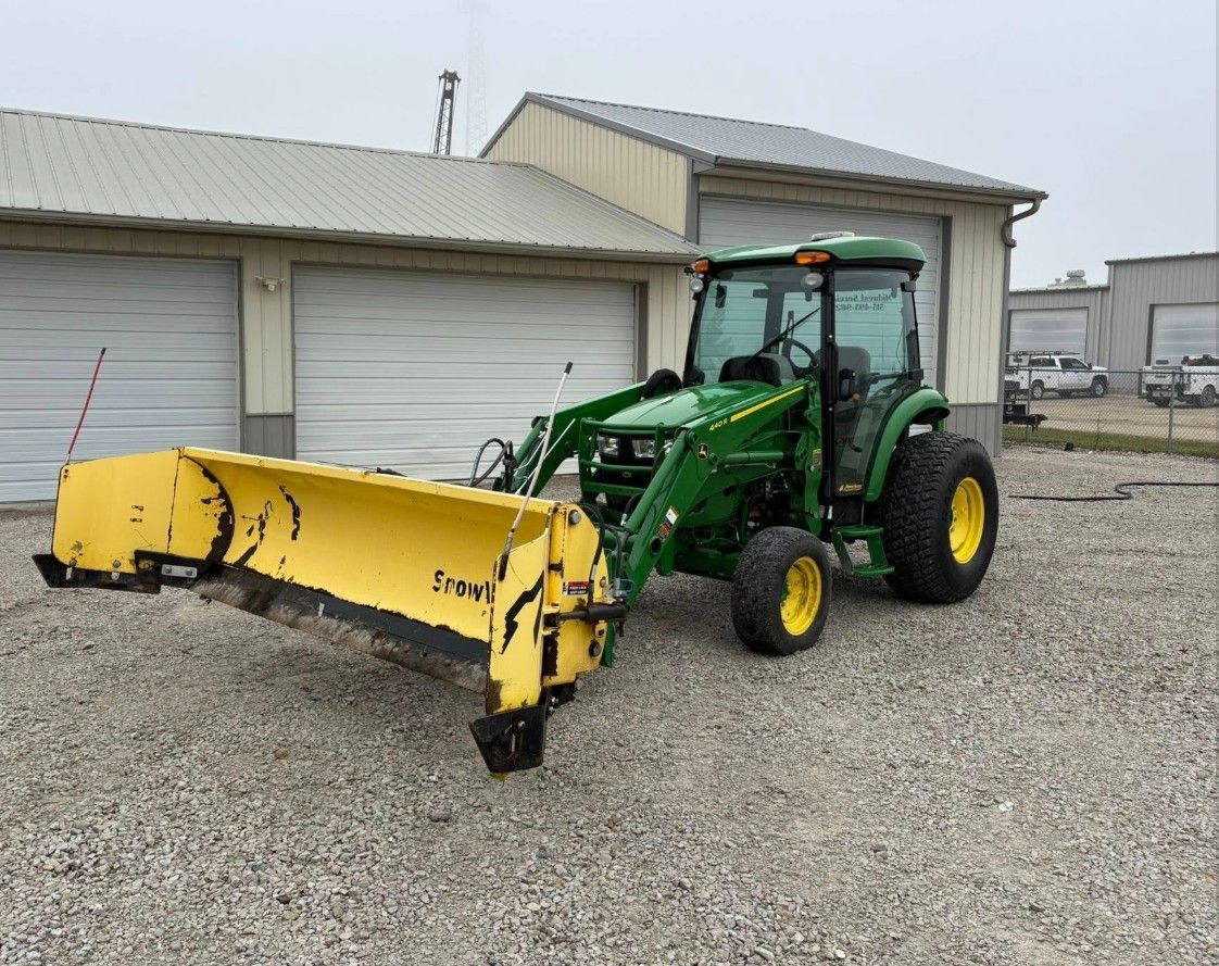 Green John Deere tractor with yellow plow in front of a building.