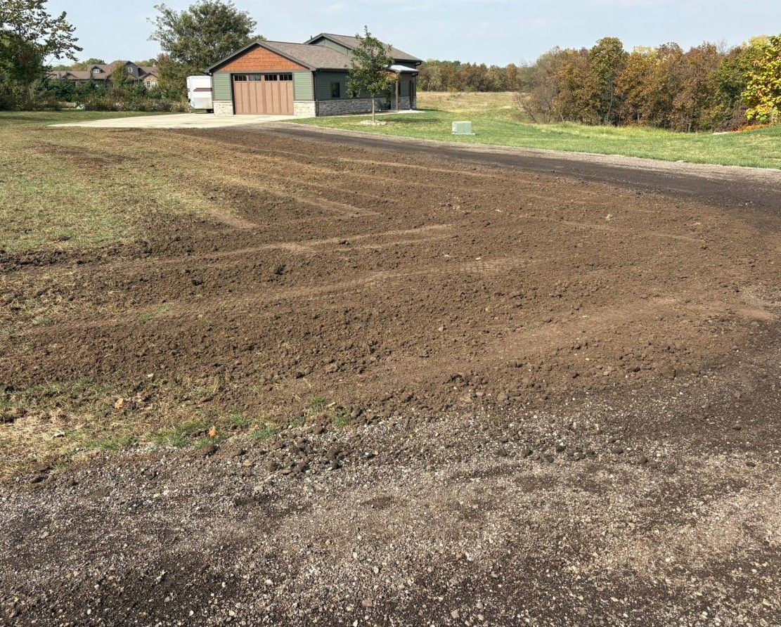Dirt field tilled and ready for planting, with a house in the background. Green grass and trees nearby.