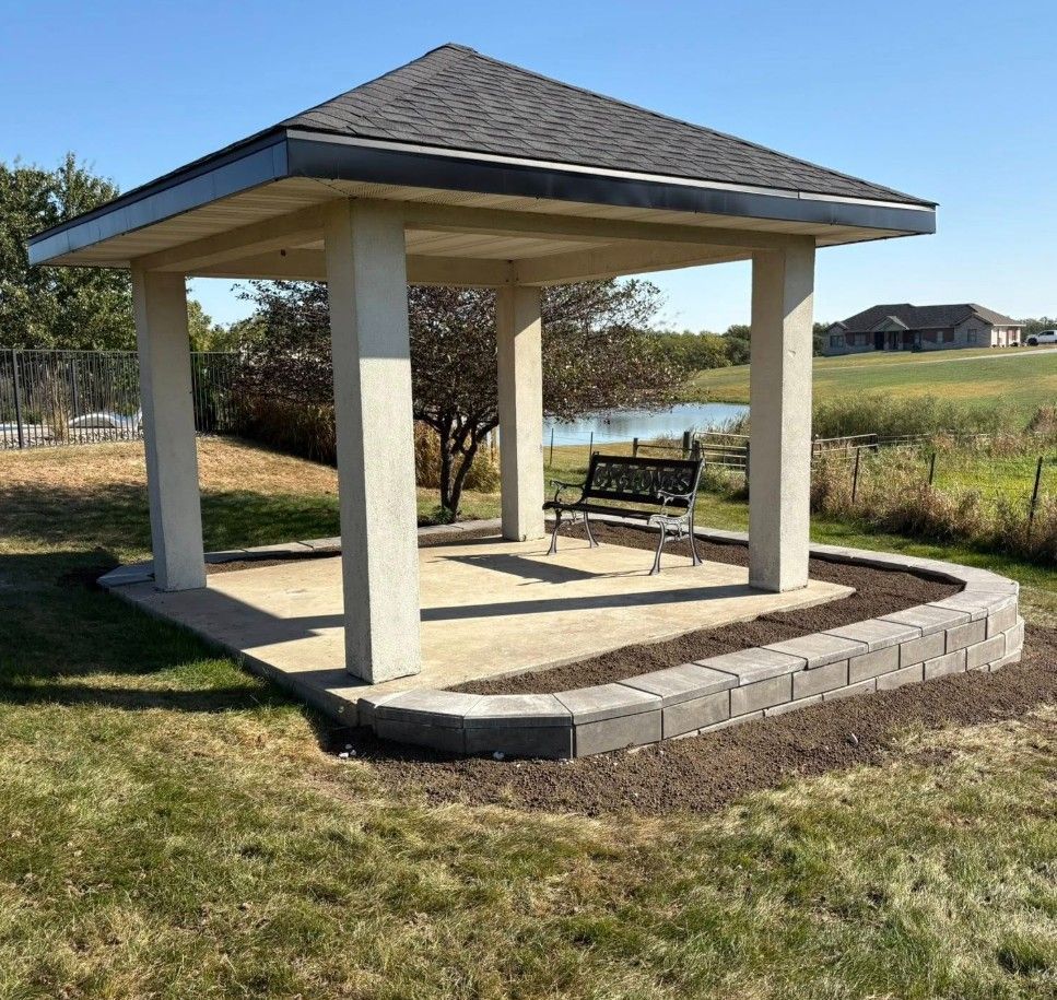 Small open gazebo with benches on a paved pad in a grassy park area