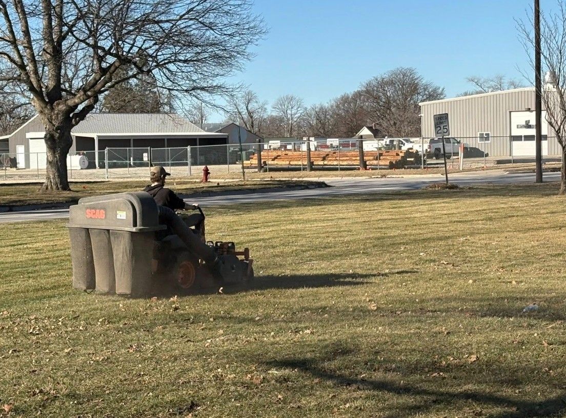 Person riding a small tractor across a grassy field near buildings on a sunny day