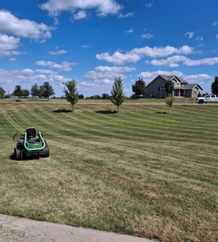 A lawn mower on a striped grassy hill with a house and blue sky in the background.
