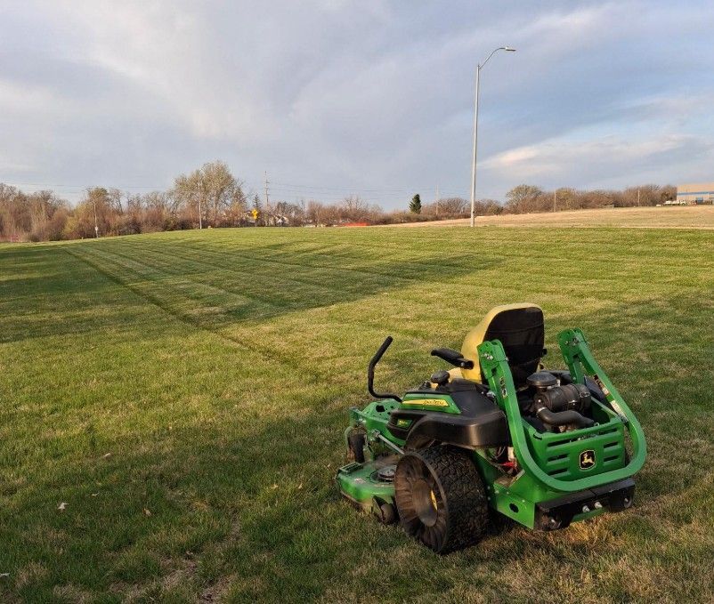 Green riding mower on a grassy hill beside a road under a cloudy sky