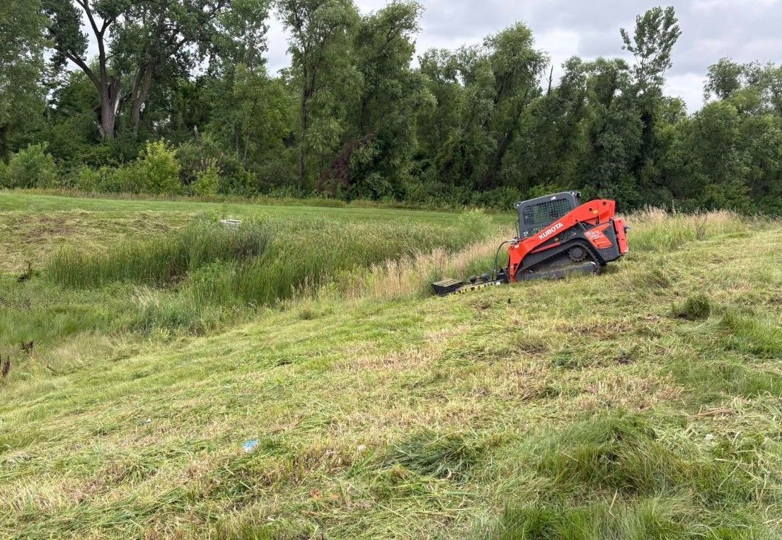 Orange excavator on a grassy slope by a tree line under an overcast sky