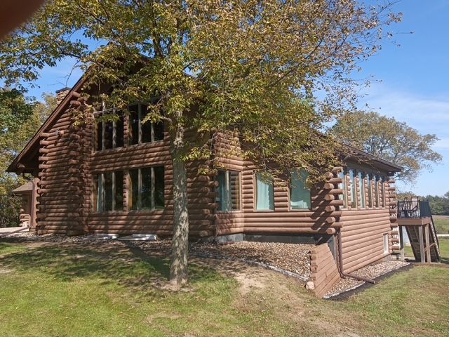 Log cabin in a sunny wooded area, with green-trimmed windows and a sloped roof.