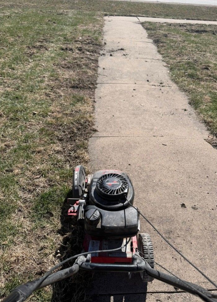 First-person view of a lawn mower being pushed along the edge of a concrete sidewalk next to a patch of grass.