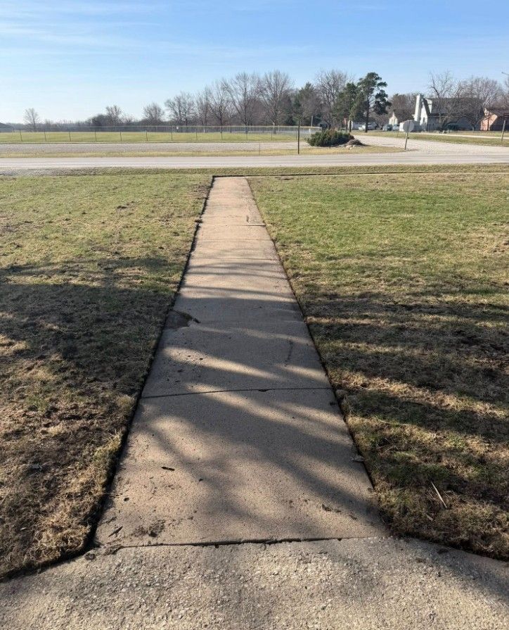 A straight concrete sidewalk stretches across a grassy lawn towards a road and trees in the distance.