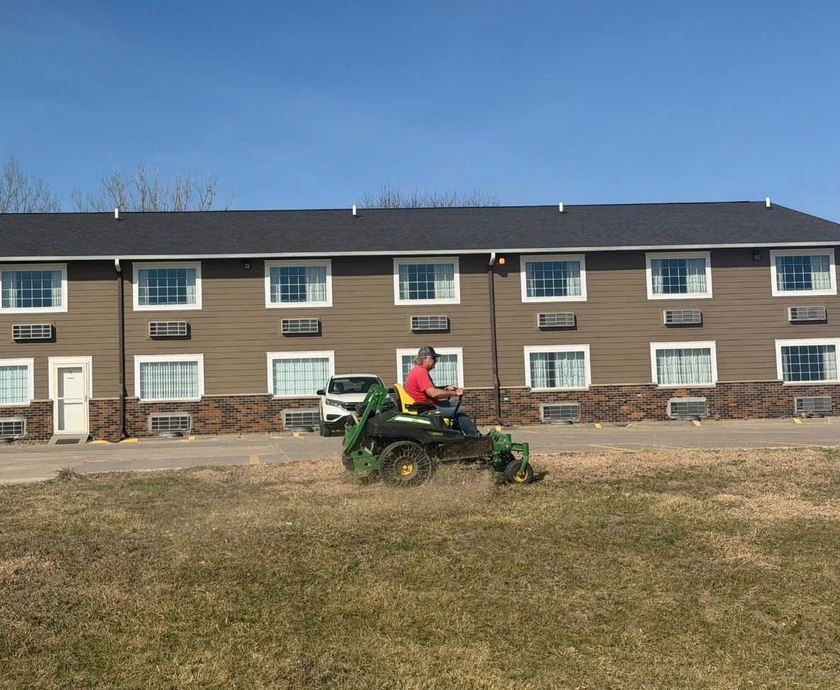 A person mows the grass in front of a tan two-story hotel under a clear blue sky.
