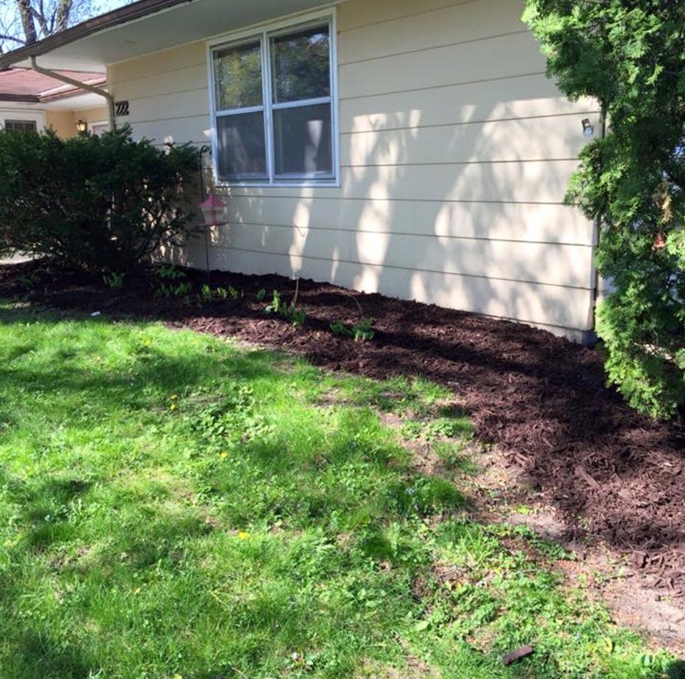 A beige house exterior with a dark mulch garden bed in front, flanked by green bushes and a lawn in the foreground.