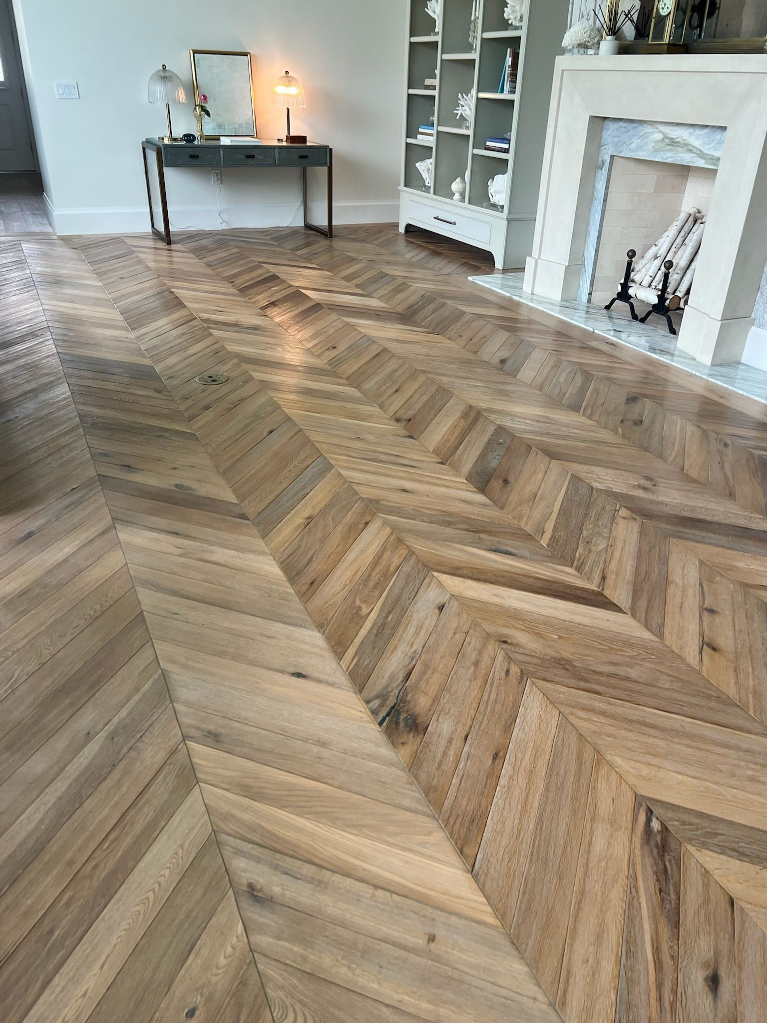 Herringbone pattern hardwood floor in a room with a fireplace, shelves, and a table with a mirror.