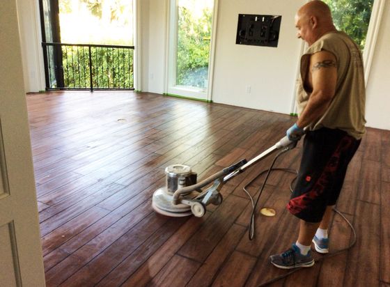 A man sanding a hardwood floor with a floor buffer in a bright room with a balcony view.