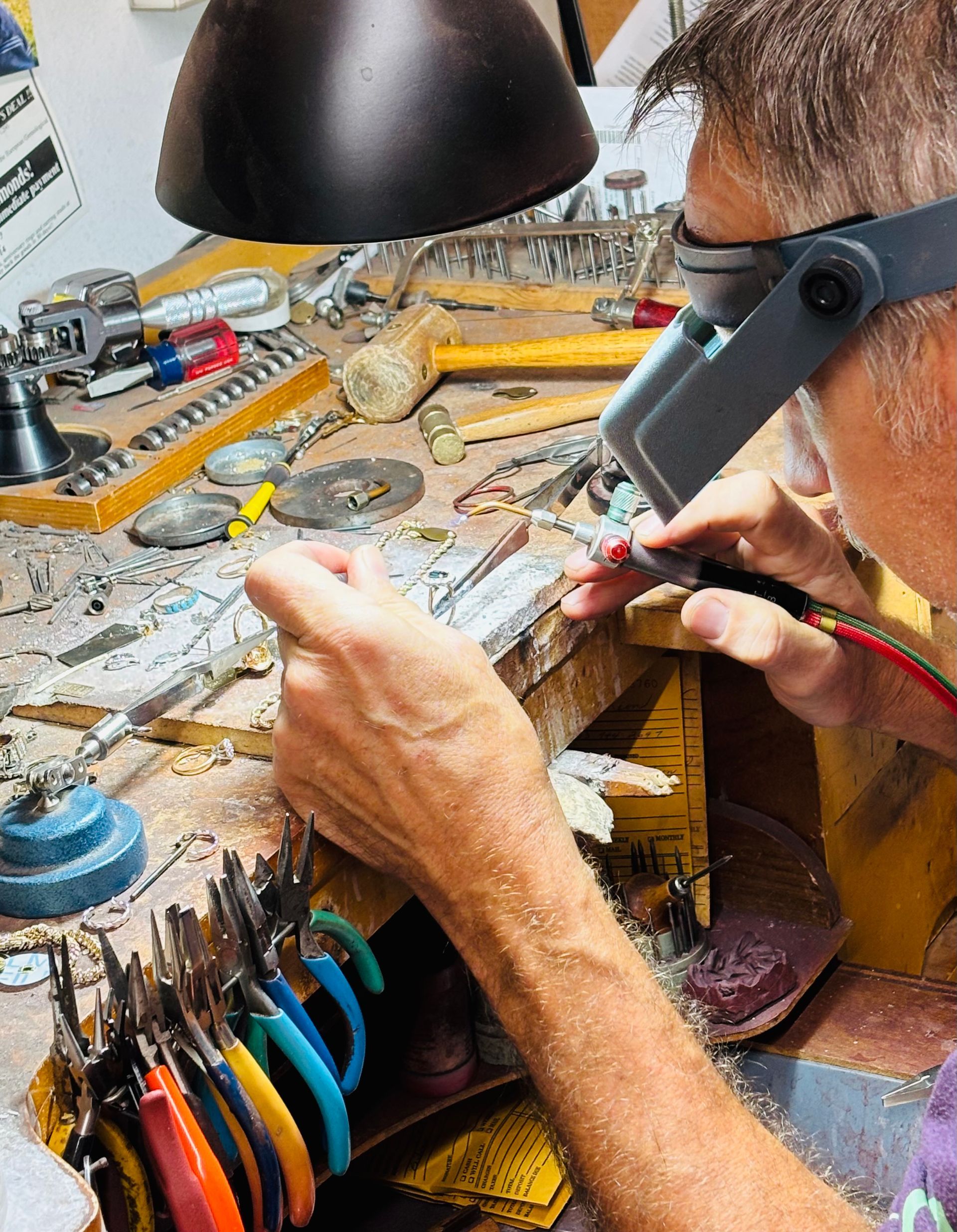 Person working on jewelry at a workbench, using tools, wearing a magnifying visor, under a lamp.