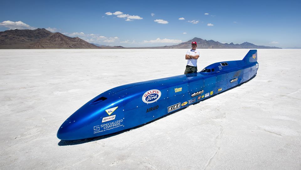 A man is standing next to a blue speed boat on a dry lake.