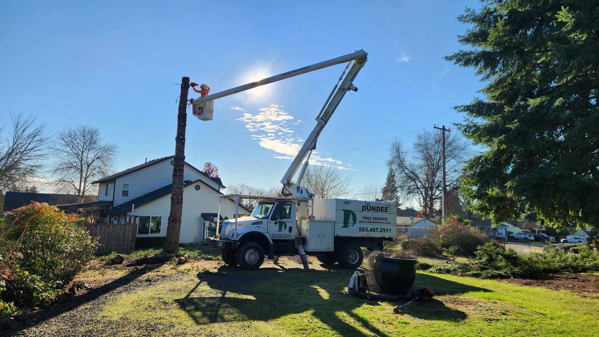 A crane truck is cutting a tree in front of a house.