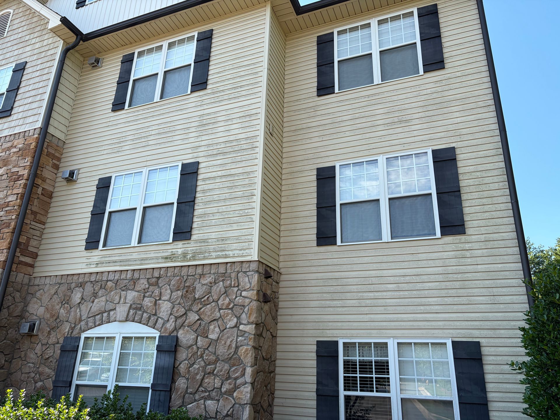 Three-story apartment building with tan siding, stone facade, and multiple windows under a blue sky
