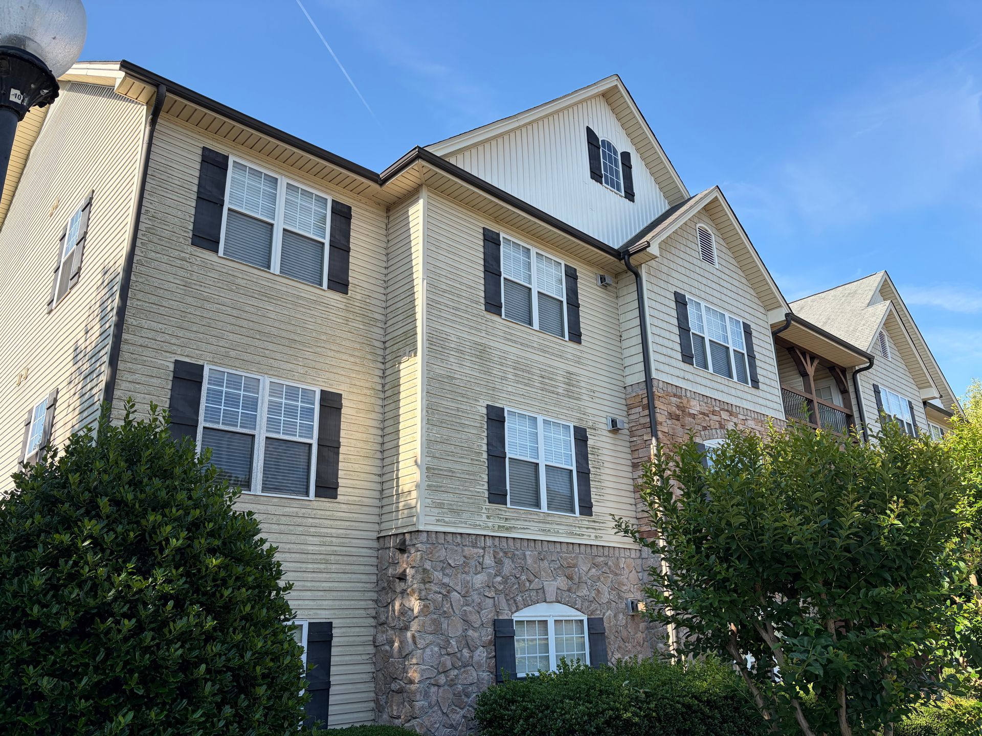 Multi-story beige brick apartment building with white-trimmed windows and green shrubs under a blue sky
