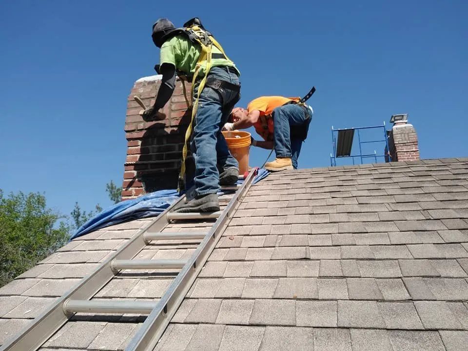 Workers repairing a shingled roof, with one on a ladder and others near a brick chimney.