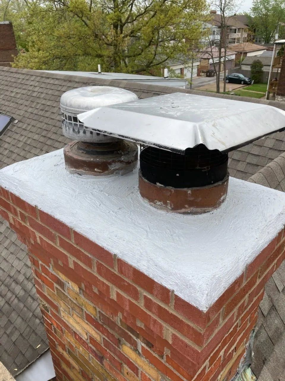 Brick chimney with two metal flue caps on a white-capped rooftop, with trees and houses in the background
