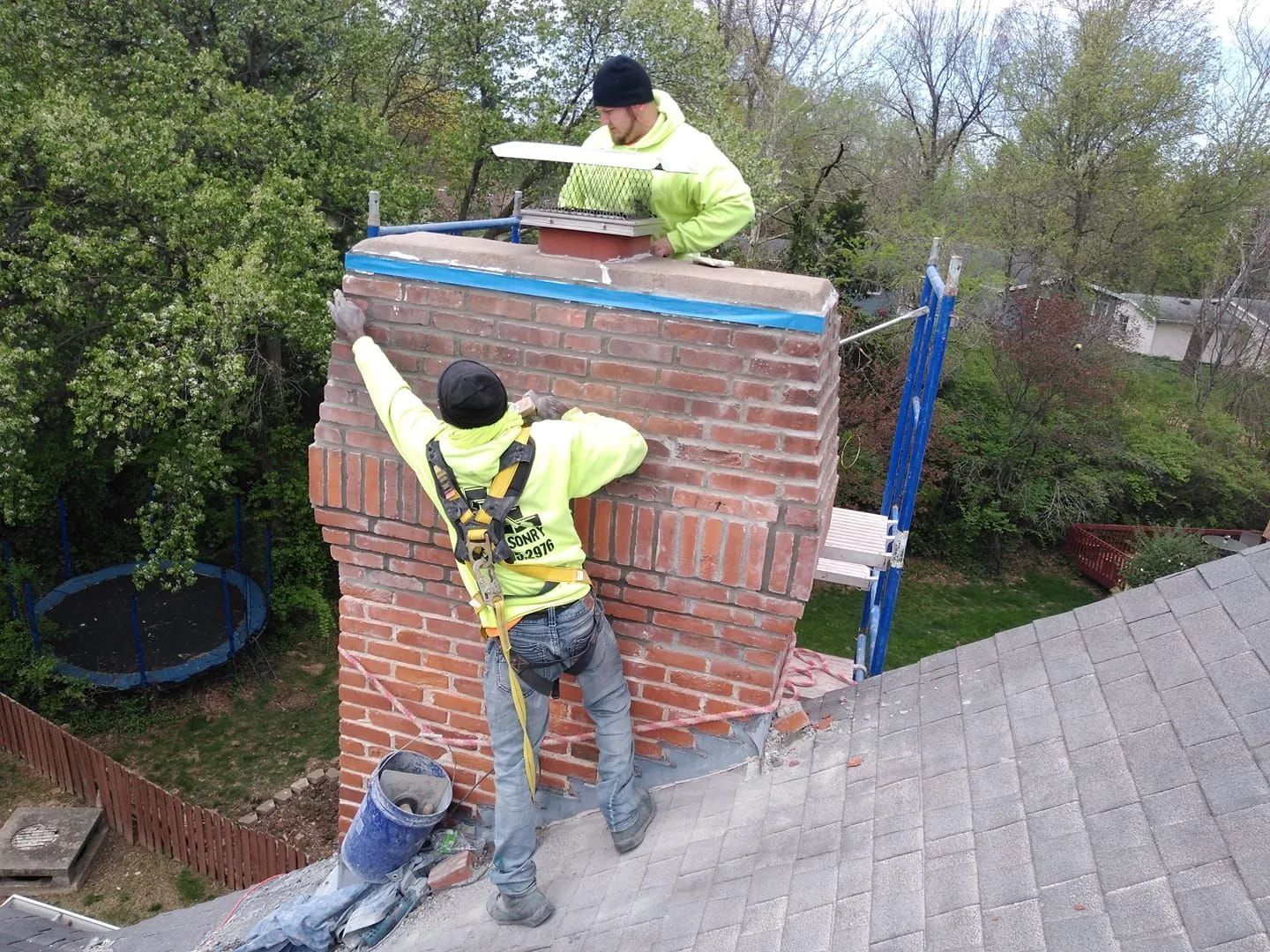 Two roofers repairing a brick chimney on a house roof, one on top and one on the side