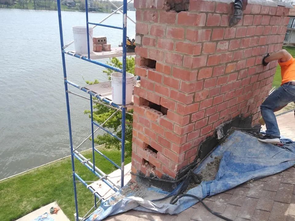 Brick structure under construction beside water, with scaffolding and a worker on a blue tarp.