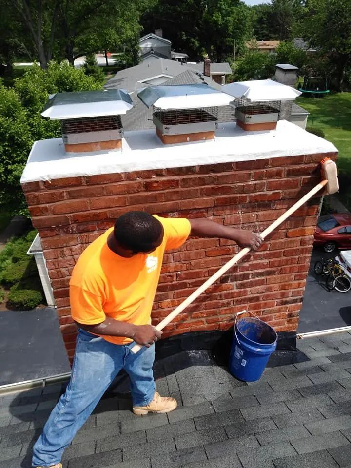 Worker power-washing a brick chimney on a rooftop, with buckets and patio below.