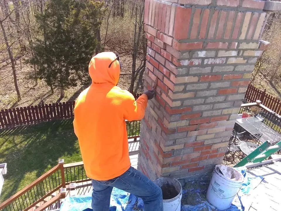 Person in bright orange jacket standing on a rooftop beside a brick chimney with paint buckets and tools nearby.