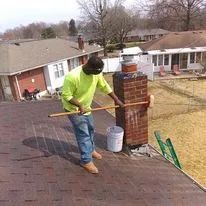 Worker pressure-washing a shingled roof beside a brick chimney, with a bucket and hose nearby.
