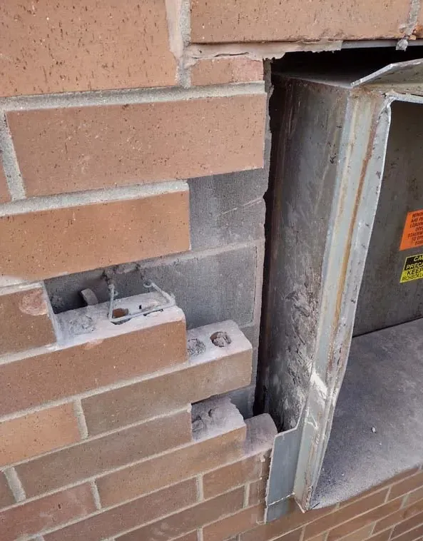 Damaged brick wall beside a metal vent or duct opening with exposed masonry and brackets