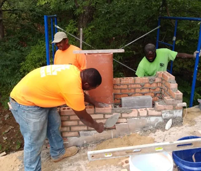 Three workers building a small brick structure outdoors, one applying mortar with a trowel.