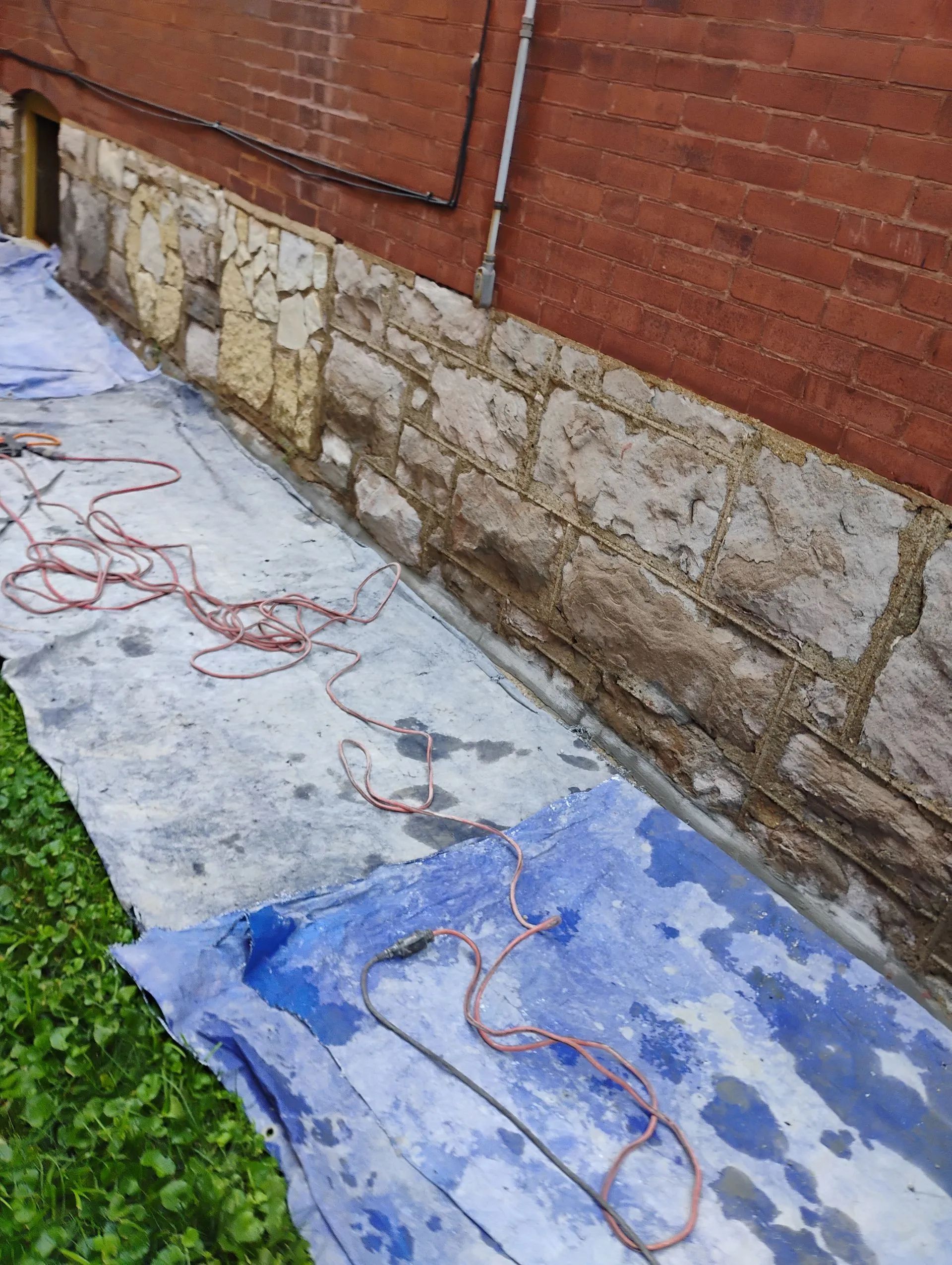 Blue tarp walkway beside a brick-and-stone wall, with coiled orange rope along the edge
