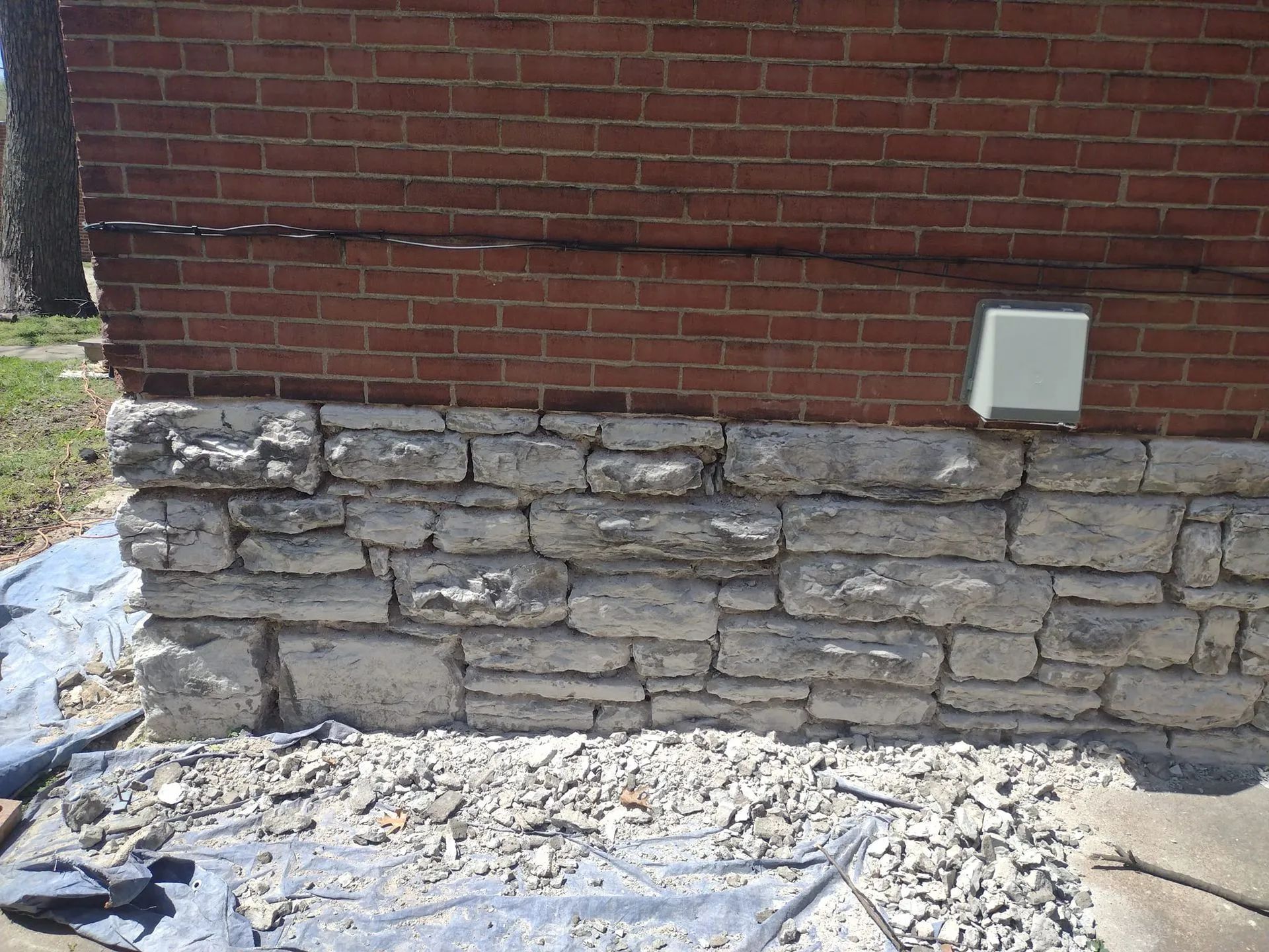 Brick wall with damaged stone foundation and rubble at the base, plus a small white utility box attached.
