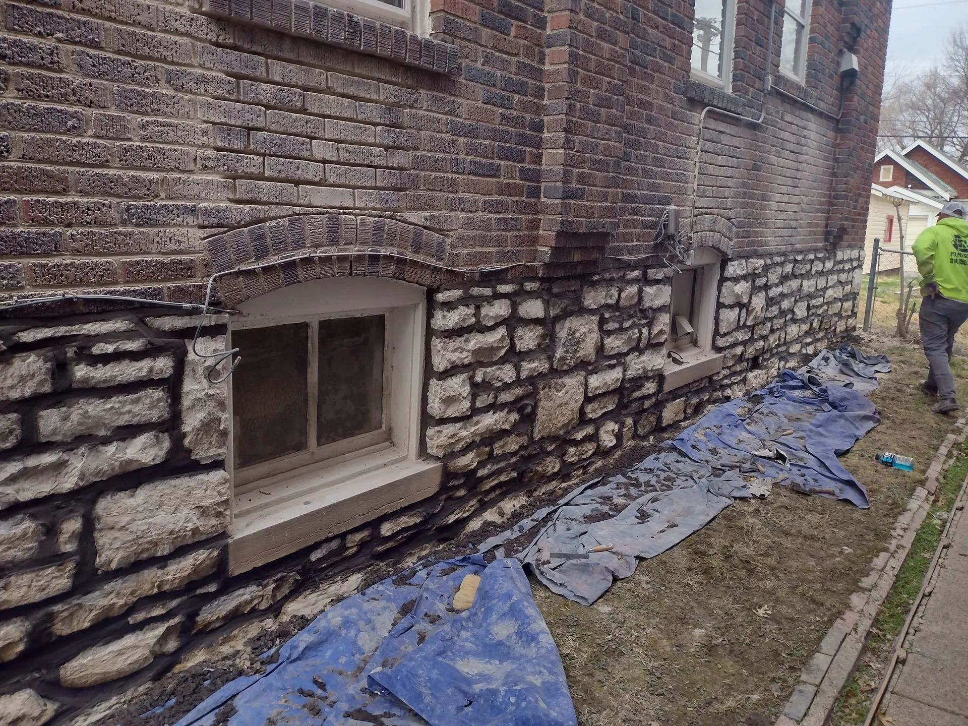 Worker repairing exterior brick wall beside a basement window, with blue tarp and tools along the ground.