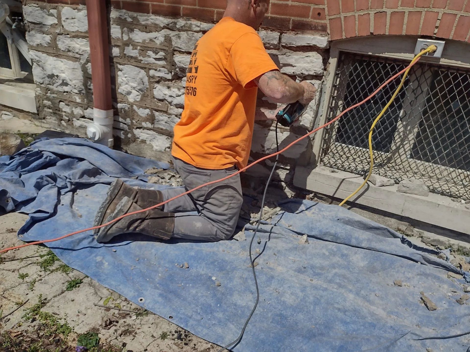 Worker in orange shirt kneels on a blue tarp, using tools near a windowed brick wall.