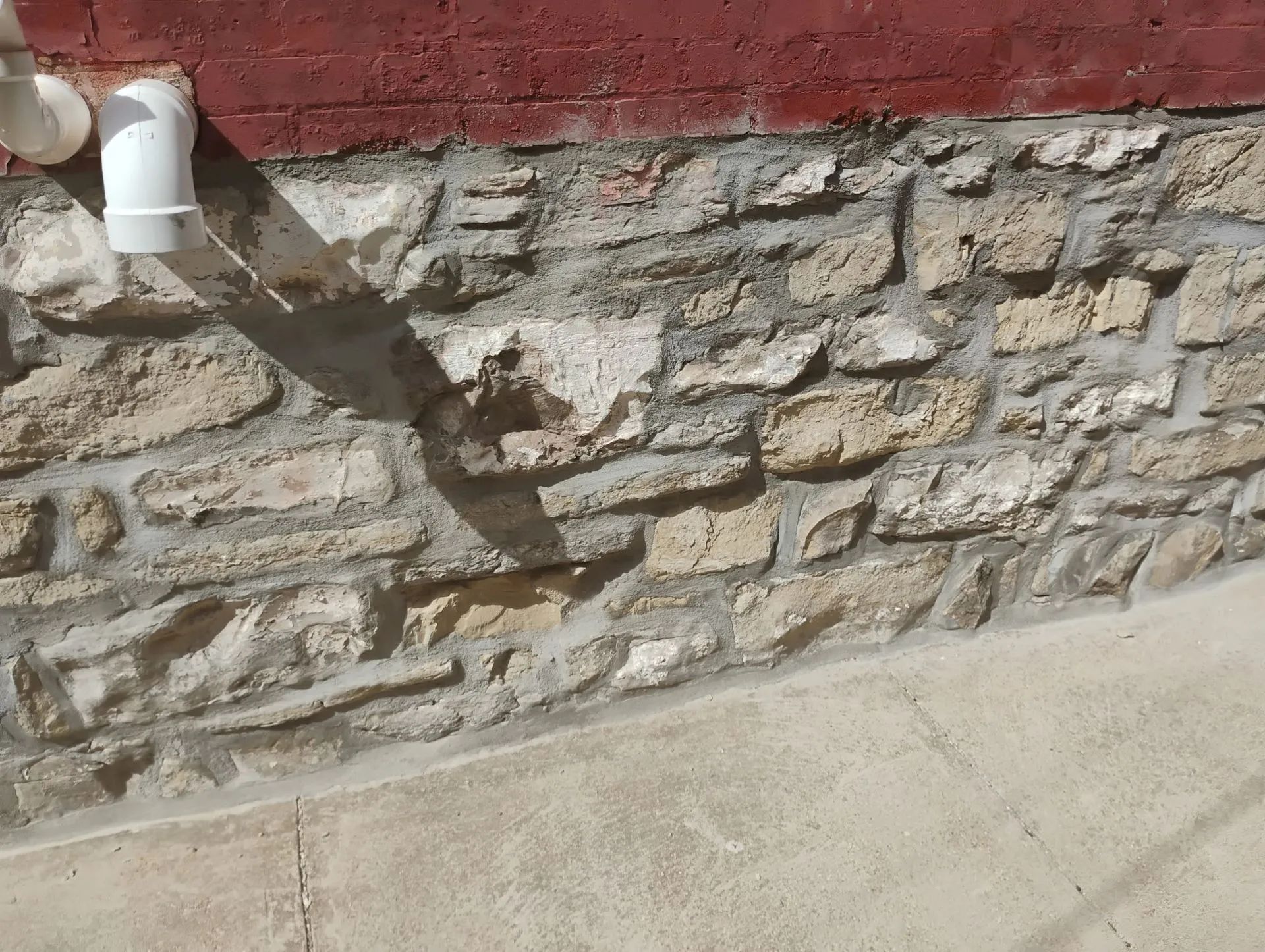 Stone wall with a white pipe casting a shadow onto a concrete sidewalk.