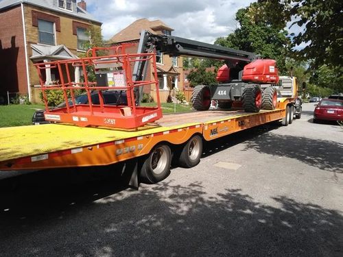 Orange flatbed trailer carrying a red boom lift parked on a residential street