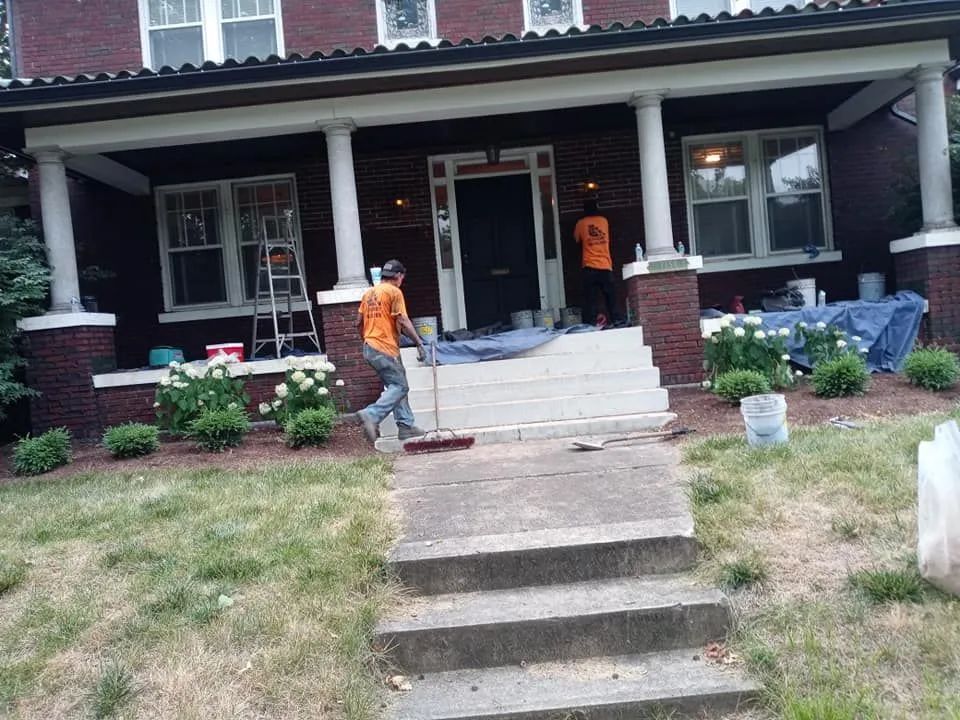 Front porch of a brick house with two workers in orange shirts and steps leading up from the yard