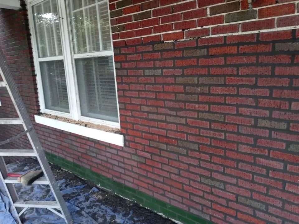 Brick exterior wall with white-trimmed window, ladder, and debris along the foundation under repair