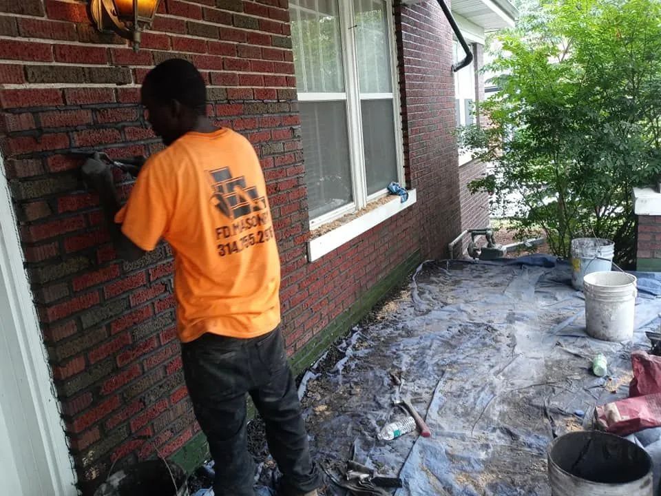 Person in orange shirt painting a brick wall beside a house on a covered patio.