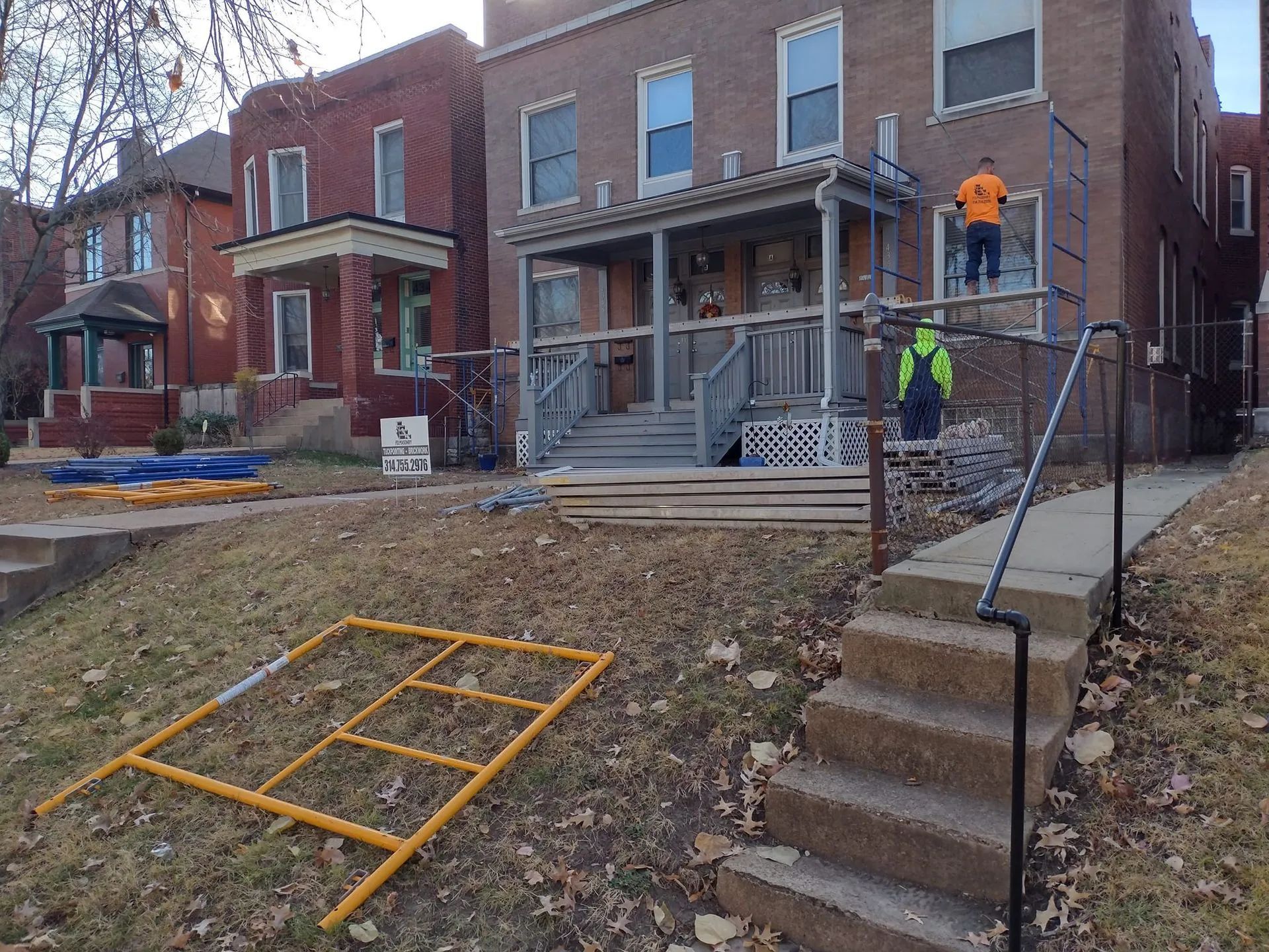 Workers on scaffolding outside a brick house under renovation, with stairs and tools in front